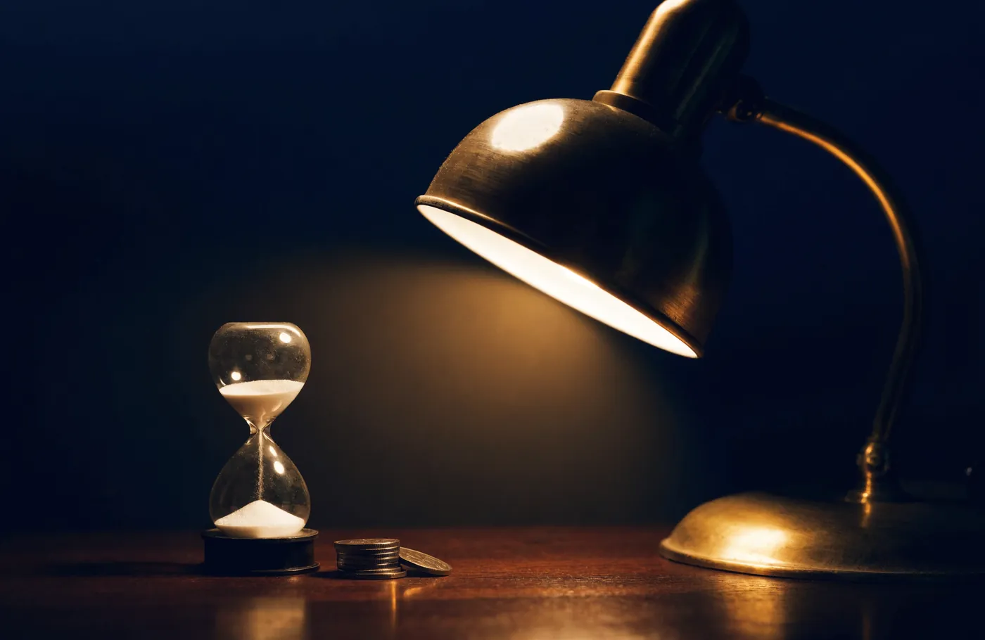 An hourglass beside stacked coins under a desk lamp