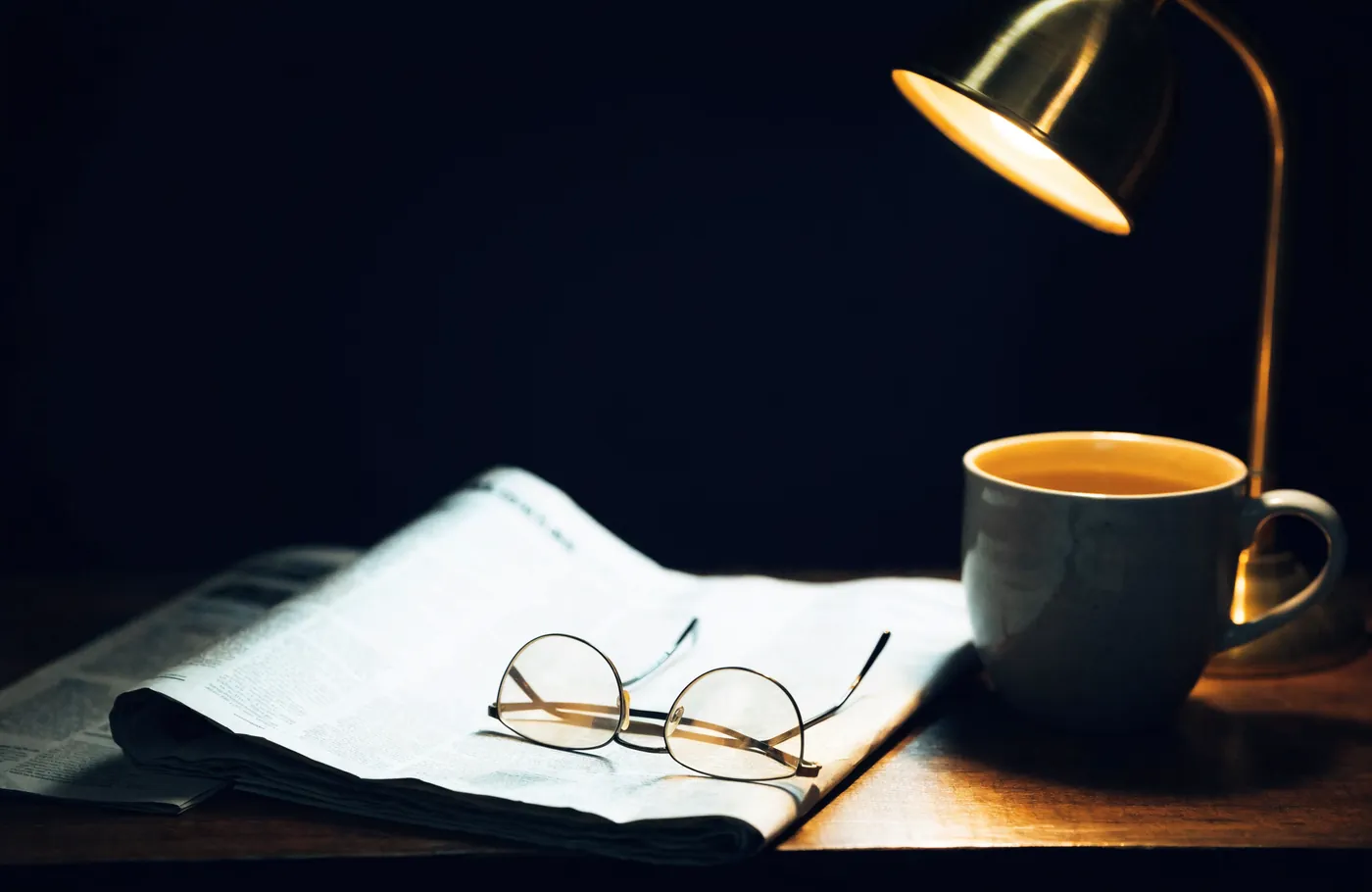 A folded newspaper and reading glasses under a desk lamp